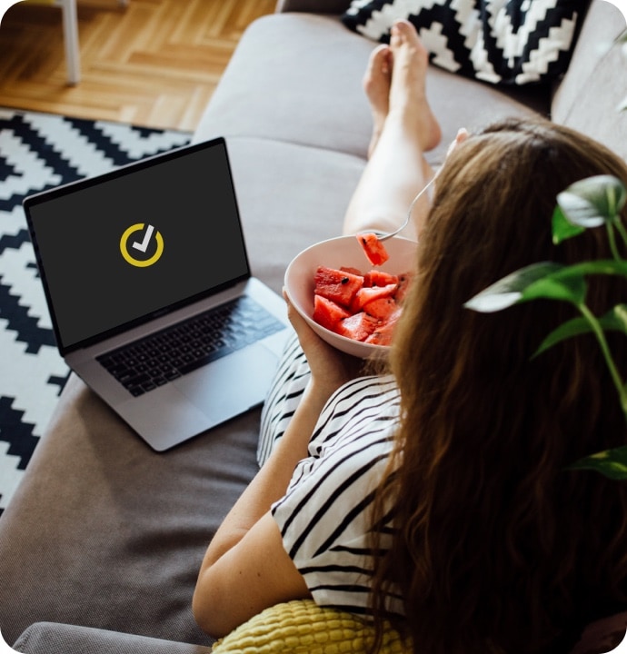Mulher deitada num sofá a comer uma tigela de fruta com um computador portátil ao lado com o logótipo Norton